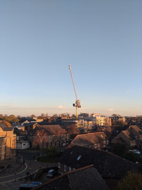 A tall construction crane with a long, extending boom is positioned above a residential area during daylight, with clear blue sky overhead and a few scattered clouds. Below the crane, multiple houses with sloped roofs and brick exteriors are visible, some with visible chimneys and small gardens. The scene includes a mixture of older and newer buildings, with the newer constructions possibly under development or renovation. In the foreground, rooftops with dark shingles are seen, with a glimpse of a street where a few parked cars and cyclists are present. The image captures the urban environment in the Alexandra Palace area, emphasizing the scale of the crane in relation to the surrounding buildings, and suggests a context of home relocation or construction activity, aligning with house removals and moving services offered by Man With a Van Bounds Green.