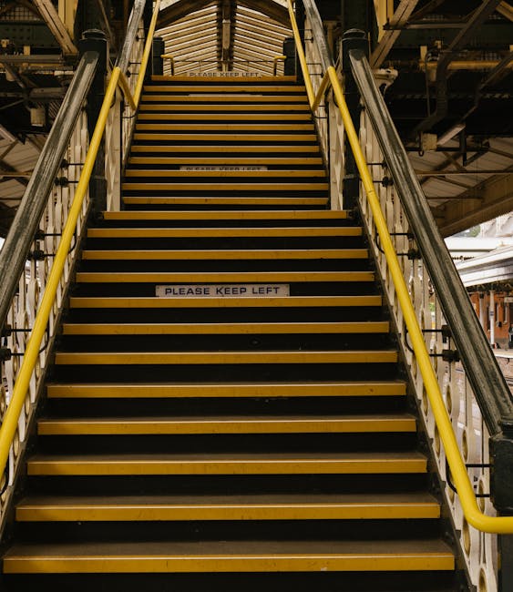A set of yellow industrial stairs with black anti-slip treads, leading upward inside a large warehouse or freight terminal. The stairs are flanked by yellow handrails on both sides, with a white sign on the middle step reading 'PLEASE KEEP LEFT'. Above the stairs, there is a metal and wooden structure supporting the ceiling, which appears to be part of the loading area infrastructure. The surrounding environment includes metal beams, piping, and industrial lighting, suggesting a busy transfer point for home relocation or furniture transport services. This image captures the technical aspect of moving logistics, illustrating the challenging access often involved in house removals in the Alexandra Palace area, where tight access solutions may be needed, as managed by companies like Man With a Van Bounds Green.