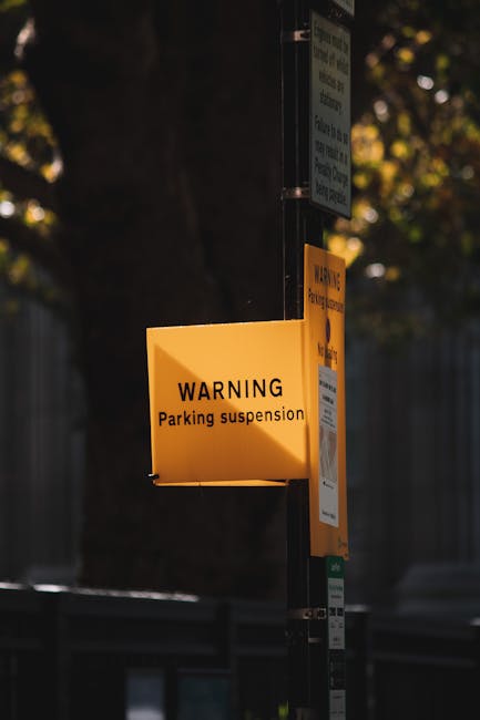 A close-up photo of a yellow warning sign attached to a metal pole, indicating a parking suspension in the vicinity of a residential property. The sign is illuminated by natural daylight, with the background showing a partially visible tree with green foliage and a blurred fence, suggesting an outdoor setting in an urban or suburban area. Behind the sign, there are other smaller notices attached to the pole, which are less clearly visible. This image captures the scene where Man With a Van Bounds Green is preparing for a home relocation, involving loading or moving furniture and boxes from a property, supporting the process of parking suspensions needed for the removal vehicle to access the premises.