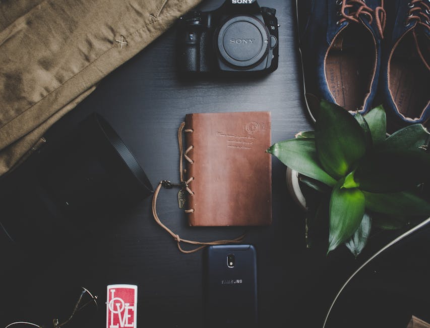 A flat-lay arrangement of various personal belongings on a dark surface, including a Sony camera with a lens cap attached, a brown leather-bound travel journal with a strap closure, a black portable speaker with a carrying strap, a Samsung smartphone, a pair of dark blue and orange sneakers, a large green leafy plant in a pot, and a neatly packed cardboard box partially covered by a beige fabric. The items are positioned in an organized manner, with some objects overlapping slightly, suggesting a scene from a home relocation or packing process. The background includes a glimpse of a door frame or doorway, indicating the items are inside an interior space, with natural lighting illuminating the scene. This detailed image captures the essentials involved in furniture transport, packing, and the moving process, consistent with the focus of Man With a Van Bounds Green in house removals.