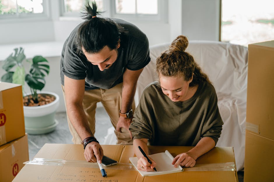 A man and a woman are engaged in a home relocation process indoors, surrounded by moving boxes made of cardboard, some sealed with clear packing tape. The woman is seated at a table, writing on a note