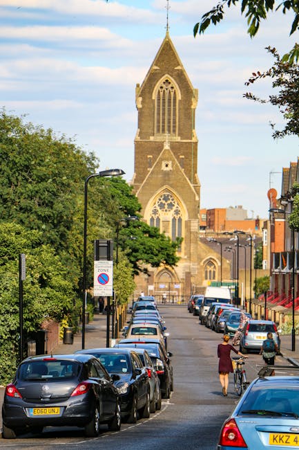 Image of a street scene on Bounds Green Road showing a row of parked cars along the curb, including a variety of hatchbacks and sedans, with some displaying UK registration plates. In the foreground, a person dressed in a dark jacket and skirt is pushing a bicycle while another individual nearby walks along the sidewalk. The street is lined with black lampposts and traffic signs, including a parking zone sign. On the right side, a building with a red awning is partially visible. In the background, there is a prominent historic church tower with pointed Gothic windows and a steeply pitched roof, framed by green trees and a clear blue sky with some clouds. This setting illustrates typical urban activity involving home relocation or furniture transport, with the church serving as a landmark near the area where Man With a Van Bounds Green offers removals and moving services, emphasizing loading and parking considerations for home moves.
