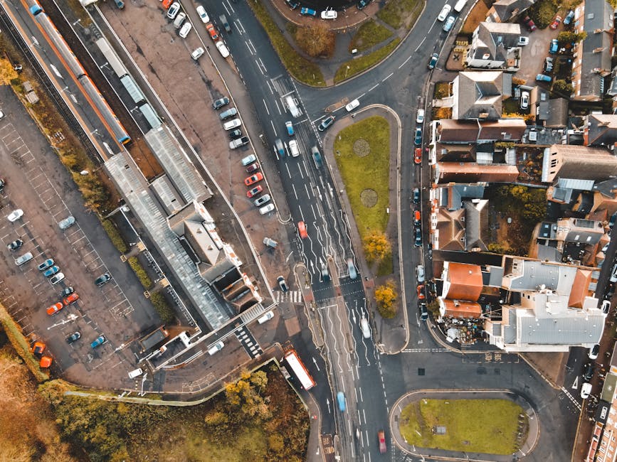 Aerial view of Bounds Green Road showing a busy urban intersection with multiple lanes of traffic, including cars, vans, and buses moving in different directions. Adjacent to the road are residential houses with pitched roofs and small gardens, as well as commercial or office buildings with glass facades and flat roofs. The scene is captured during daylight with overcast skies, and there are designated parking areas with cars parked side by side along the streets and in nearby lots. A metal and glass pedestrian footbridge crosses over the road, facilitating safe crossing for pedestrians. Green patches with trees and small grassy areas are visible near the road's intersections, indicating landscaped urban spaces. This setting relates to home relocation and furniture transport activities as part of professional removals services, such as those provided by Man With a Van Bounds Green, involved in packing, loading, and transporting household items through busy city streets.