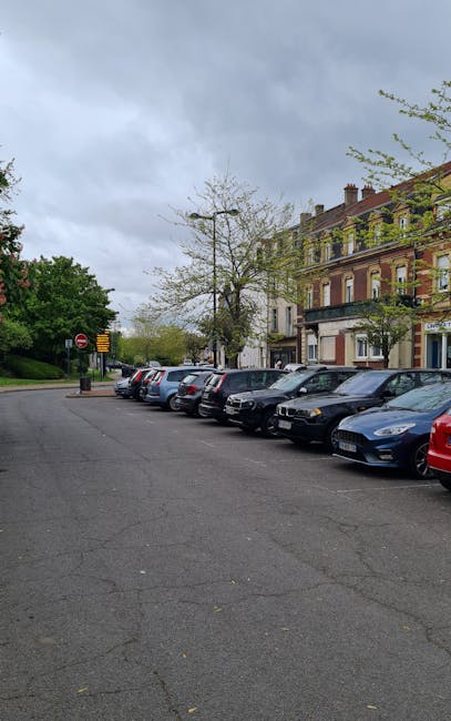 Image of a street scene on Bounds Green Road showing a row of parked cars along the curb, including a variety of hatchbacks and sedans, with some displaying UK registration plates. In the foreground, a person dressed in a dark jacket and skirt is pushing a bicycle while another individual nearby walks along the sidewalk. The street is lined with black lampposts and traffic signs, including a parking zone sign. On the right side, a building with a red awning is partially visible. In the background, there is a prominent historic church tower with pointed Gothic windows and a steeply pitched roof, framed by green trees and a clear blue sky with some clouds. This setting illustrates typical urban activity involving home relocation or furniture transport, with the church serving as a landmark near the area where Man With a Van Bounds Green offers removals and moving services, emphasizing loading and parking considerations for home moves.