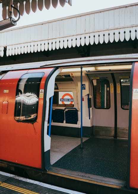 The open doors of a London Underground train at Bounds Green station reveal the interior with two blue upholstered seats, grab handles, and safety signage. The train is red with a white interior and is parked on the platform beneath a white, curved canopy with hanging decorative elements. Outside, the platform edge is visible, indicating a site prepared for home relocation or furniture transport as part of a house move. This setting captures the typical station environment where moving logistics, such as loading boxes and furniture, may take place with assistance from a professional removals service like Man With a Van Bounds Green, supporting efficient packing and moving processes.