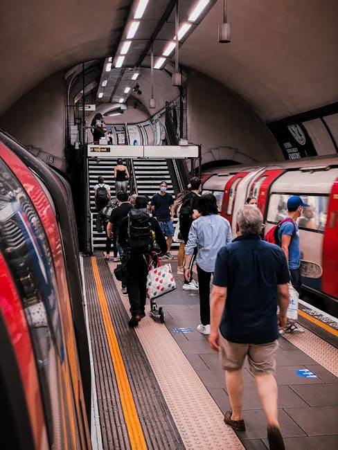 A busy underground train station platform with passengers walking along the edge near parked London Underground trains. The scene shows a diverse group of people, some carrying backpacks and suitcases, wearing casual clothing and face masks. The platform features tactile paving strips and safety lines near the trains, with stairs and a mezzanine level visible in the background, leading to an upper exit area. Overhead lighting illuminates the curved tunnel-style ceiling. This setting relates to home relocation or furniture transport logistics, which are often part of house removals services provided by Man With a Van Bounds Green, especially during moving processes involving travel between properties or stations.
