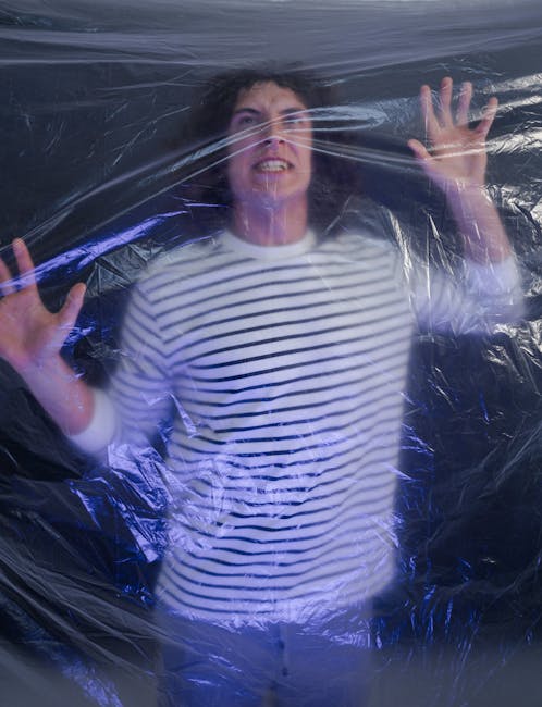 A woman wearing a white and navy striped long-sleeve shirt is positioned behind a transparent plastic sheet, with her face partially pressed against it and her hands raised, pressing against the plastic. The background appears to be a dark, possibly indoor environment, with reflections and lighting creating a slightly glossy effect on the plastic. The scene suggests the plastic sheet is being used as a protective barrier, potentially as part of a packing or moving process. This image is associated with house removals and furniture transport services offered by Man With a Van Bounds Green, highlighting careful handling and protection of belongings during home relocation or bulky waste removal in the Bounds Green area.