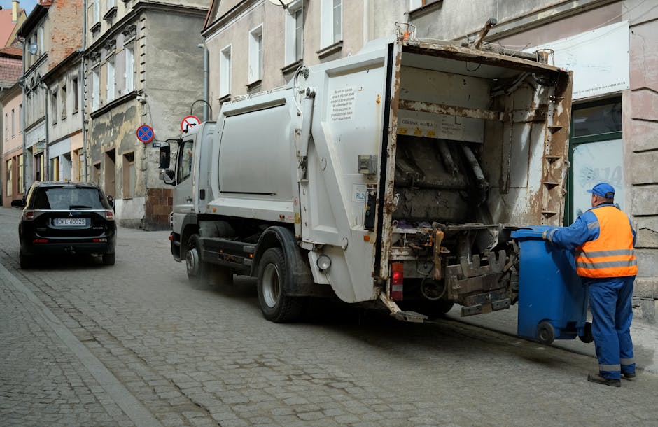 A street scene in Bounds Green showing a white garbage collection truck operated by Man With a Van Bounds Green with its rear open, ready to collect bulky waste. A worker wearing a blue uniform and orange high-visibility vest is in the process of emptying a blue wheeled waste bin into the truck’s compactor area. The truck is parked parallel to the pavement, with a black car parked further along the street. The surrounding environment includes a row of older mixed-use buildings with various window styles, some with old paintwork and weathered facades. The scene is lit by daylight, with overcast skies resulting in diffuse lighting. The image captures the logistics involved in home relocation waste disposal and the process of loading waste materials such as large household items, packaged boxes, or bulky furniture into the vehicle for removal, reflecting the efficient hauling services offered by [COMPANY_NAME].