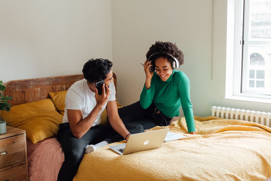 A young man and woman are seated on a bed inside a room with natural light coming through a large window. The man, dressed in a white t-shirt and dark trousers, is holding a smartphone to his ear whil