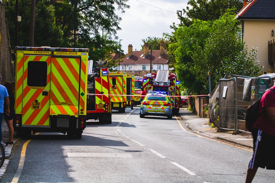 Multiple emergency service vehicles, including a London ambulance and several fire trucks with bright yellow and red chevron markings, are positioned along a residential street in Bounds Green (N11). The scene shows the vehicles blocking the road, with some parked on the pavement, indicating a response to an incident. A police car with flashing blue lights is also present, parked behind the fire trucks, while the road is cordoned off with red and white tape. On either side of the street, there are houses, trees, and scaffolding covered with protective fabric, suggesting ongoing safety or construction work. Two pedestrians are visible, one on each side of the street, observing the scene. The environment is well-lit with natural daylight, and the background features a row of houses with chimneys and rooftops. This setting illustrates a coordinated emergency response, potentially involving rescue and safety operations during a home relocation or major incident in the area, with Man With a Van Bounds Green providing professional removals and logistics support.