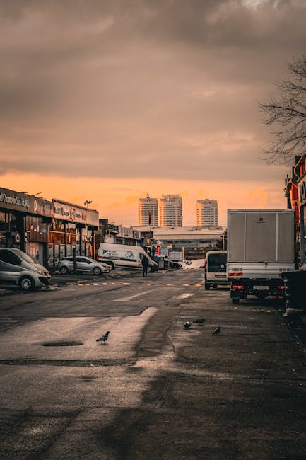 A wide view of a urban commercial area during sunset, featuring a partly cloudy sky with orange hues. In the foreground, a paved parking lot contains several vehicles, including a white delivery van positioned near a loading area, and a small grey car parked to the left. A man in dark clothing is seen walking between the vehicles, likely involved in a moving or loading process. On the right side, a large white box truck is parked close to a building with a red brick facade, which appears to be part of a shop or warehouse. On the left side, another vehicle is partially visible, parked adjacent to storefronts with signage. The scene captures the environment typical of a home or commercial relocation, with movement and parking indicative of a furniture transport or packing and moving activity, supported by Man With a Van Bounds Green for removals services.