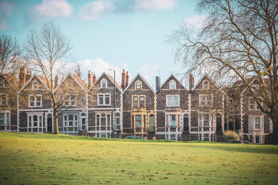 A row of Victorian-style terraced houses constructed from grey stone with white window frames and decorative elements, situated along a quiet residential street. The front gardens are enclosed by low fences, and there's a small pathway leading to each entrance. Tall, leafless trees flank the houses on either side, indicating an autumn or winter season. In the foreground, there is a wide, well-maintained grassy area, possibly a park or communal space, with a few patches of fallen leaves scattered across the grass. The sky is partly cloudy with patches of blue visible, and the lighting suggests a clear, daytime setting. The scene reflects a typical London suburb environment, which is relevant to house removals and home relocation services like those offered by [COMPANY_NAME], illustrating the context of residential property transfers, packing, and furniture transport involved in a home move in N11, BOUNDS GREEN. This imagery supports understanding of moving logistics in the area, aligning with guidance on obtaining council permits for removals in the district.