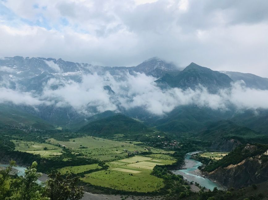 The image depicts a panoramic view of a lush green valley with patches of farmland, bordered by a winding river flowing through the landscape. Towering mountains in the background are partially obscured by low-hanging clouds and mist, creating a dramatic atmosphere. The terrain includes steep slopes and rocky outcrops, with dense forested areas visible at the base of the mountains. The natural environment appears remote and pristine, characteristic of a mountain or countryside setting. This scene could be relevant in the context of home relocation or moving logistics, illustrating the various terrains that may be navigated during furniture transport or packing and moving processes, especially in regions like Haringey or similar rural areas. The image is well-lit with natural daylight, emphasizing the vibrant greenery and rugged mountain features, which are typical of scenic outdoor landscapes often encountered during detailed house removals.
