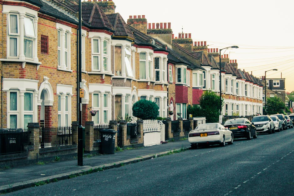 A row of Victorian-style terraced houses constructed from grey stone with white window frames and decorative elements, situated along a quiet residential street. The front gardens are enclosed by low fences, and there's a small pathway leading to each entrance. Tall, leafless trees flank the houses on either side, indicating an autumn or winter season. In the foreground, there is a wide, well-maintained grassy area, possibly a park or communal space, with a few patches of fallen leaves scattered across the grass. The sky is partly cloudy with patches of blue visible, and the lighting suggests a clear, daytime setting. The scene reflects a typical London suburb environment, which is relevant to house removals and home relocation services like those offered by [COMPANY_NAME], illustrating the context of residential property transfers, packing, and furniture transport involved in a home move in N11, BOUNDS GREEN. This imagery supports understanding of moving logistics in the area, aligning with guidance on obtaining council permits for removals in the district.