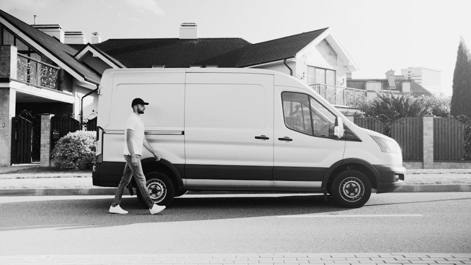 A professional mover from Man With a Van Bounds Green is seen loading cardboard boxes and packed items into a white panel van outside a modern building during daylight hours. The mover, dressed in dar
