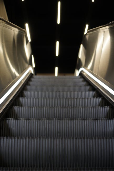 A close-up view from the bottom of an upward-moving escalator with metal handrails and metallic steps, illuminated by bright, evenly spaced linear lights on the ceiling above. The escalator is situated indoors, possibly within a modern shopping centre or transport hub, with the surrounding environment featuring dark walls and a sleek, contemporary design. This scene captures the process of home relocation or furniture transport as part of a moving or packing operation, with the focus on the systematic movement of objects, although no furniture or boxes are visible in this specific image, the setting suggests a well-organized loading or unloading area for house removals conducted by Man With a Van Bounds Green, aligned with the topic of loading processes and move-day logistics.
