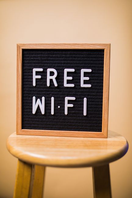 A close-up of a wooden-framed letter board placed on a small, round wooden stool. The board has a black background with white plastic letters spelling out 'FREE WIFI'. The setting appears to be indoors with a beige wall in the background. The stool's legs are visible, and the lighting is even, highlighting the sign's message. This image relates to home or business environments where free Wi-Fi access is offered, which can be relevant for customers during house removals or relocation visits facilitated by Man With a Van Bounds Green, a professional removals service within the context of moving and packing logistics.