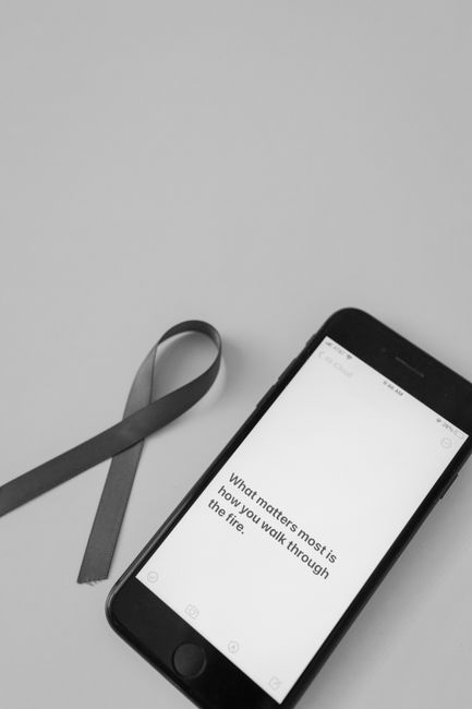A smartphone displaying a motivational quote about perseverance on a plain, light-colored surface next to a dark grey fabric strap. The phone screen shows the text: 'What matters most is how you walk through the fire.' The background is empty, with no other objects visible, creating a clean and neutral setting that emphasizes the device and the strap, which may be used for carrying or securing items during a home relocation or furniture transport process. This image illustrates the importance of careful handling and preparation in moving logistics, as seen in professional removals services like those offered by Man With a Van Bounds Green.