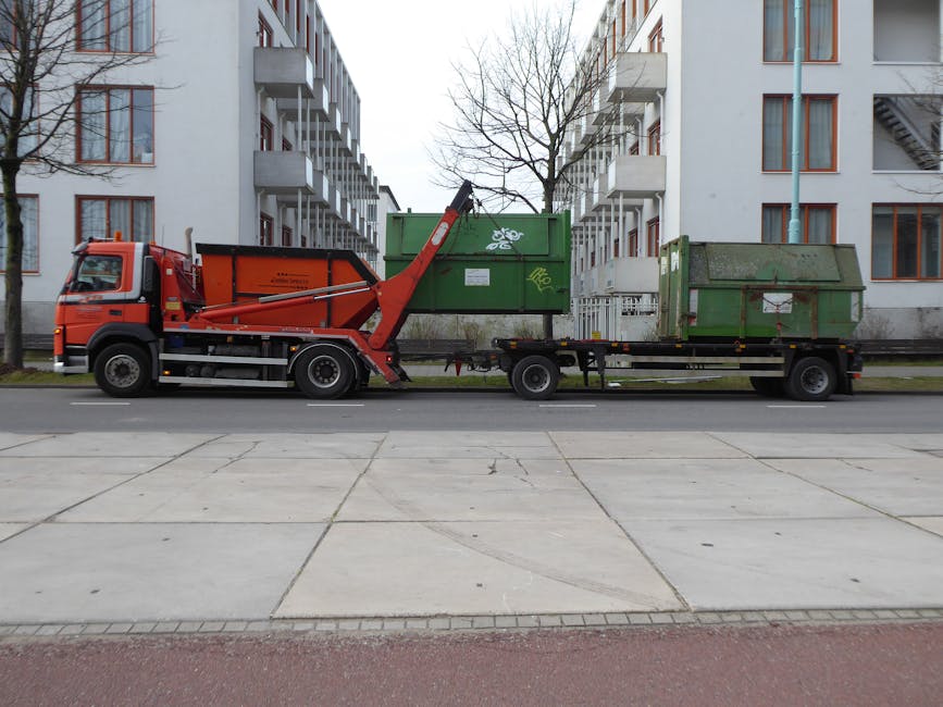 A red and black flatbed truck with a mechanical crane arm attached is parked on a street in Bounds Green, Haringey, during daytime. The truck is operated by Man With a Van Bounds Green, part of a house removals service, and is in the process of loading or unloading two large, green metal waste containers positioned on the flatbed. The containers appear to be used for disposal or waste management, with one showing graffiti or markings on its surface. The truck is situated adjacent to a modern residential building with white walls, multiple balconies, and large windows, and is parked on a paved area with trees and a lamppost visible in the background. The scene captures a typical outdoor setting for furniture transport or packing and moving activities, focusing on loading logistics within an urban environment.