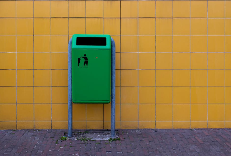 A bright green public waste bin with a black icon of a person disposing of rubbish, mounted on two metal posts in front of a yellow tiled wall on a pavement. The bin is positioned at an accessible height with a rectangular opening at the top for waste disposal. The surrounding paving consists of red bricks, and there are small patches of grass and dirt at the base of the posts. This image captures the outdoor environment where house removals and packing are prepared, highlighting logistical elements involved in home relocation, such as waste disposal. The presence of the bin and the tiled wall suggests a residential or commercial area near a property where furniture transport and packing materials might be disposed of during a move. Man With a Van Bounds Green offers removal services that facilitate moving processes like these, ensuring proper disposal and transportation of household items.