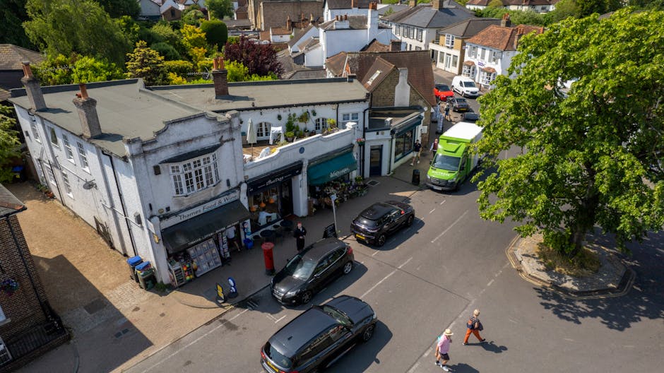 An aerial view of a busy street in Bounds Green showing a white two-storey building with shopfronts and awnings, surrounded by parked vehicles including a black car, a silver car, and a green van, with several pedestrians walking on the pavement. To the right, a large green tree provides shade, while a man with a pink hat and orange trousers is walking across the road. In the background, residential houses with pitched roofs are visible, along with greenery and trees. The scene depicts a typical urban environment suitable for house removals and furniture transport, with vehicles positioned for loading and unloading, and some items possibly being prepared for a home relocation process, which Man With a Van Bounds Green offers as part of their removals services.